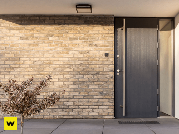 Moderne Haustür aus dunklem Aluminium neben heller Klinkerfassade und kleinem Strauch.